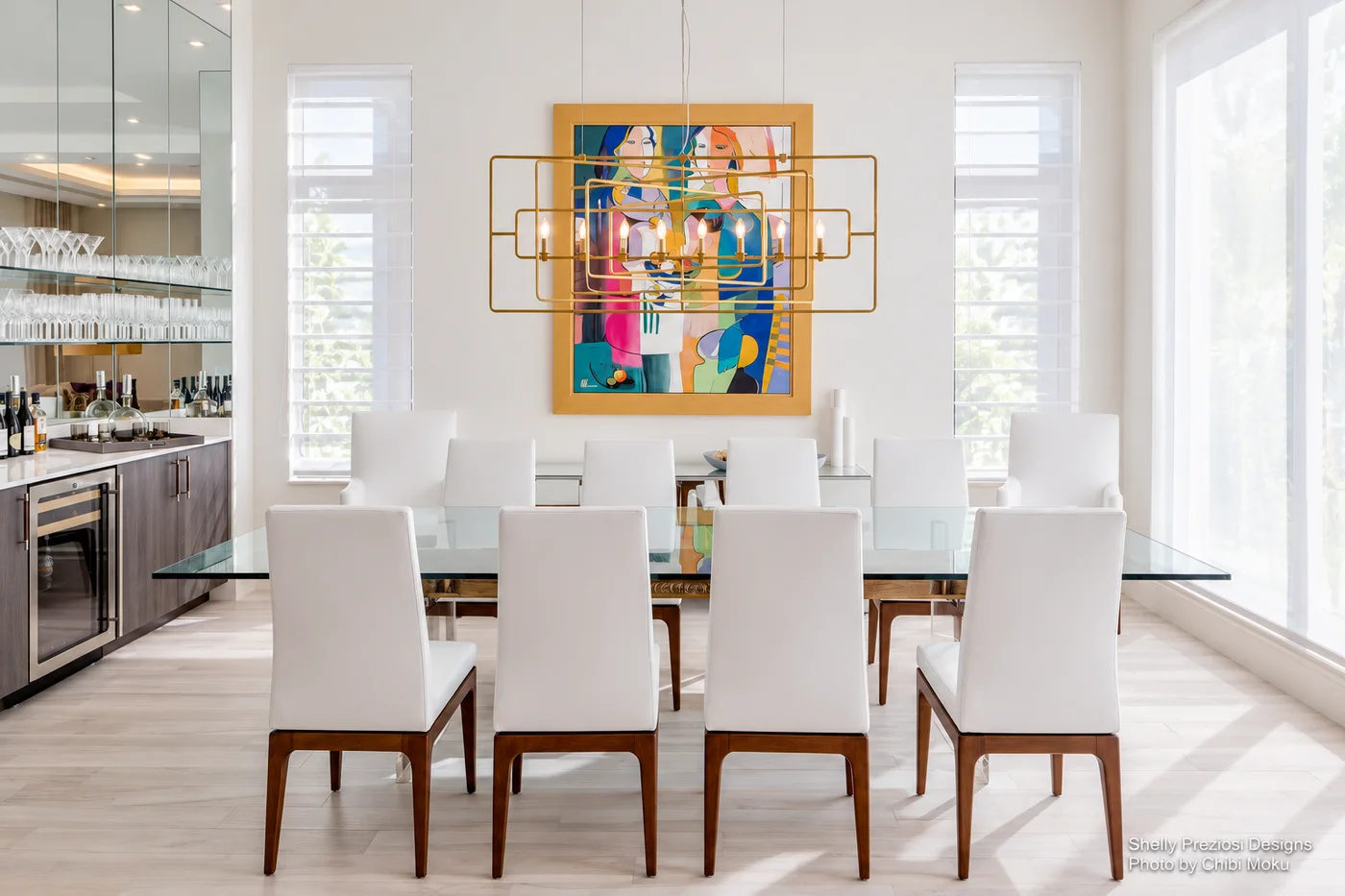 A bright dining room with white chairs, colorful artwork, and a geometric gold chandelier